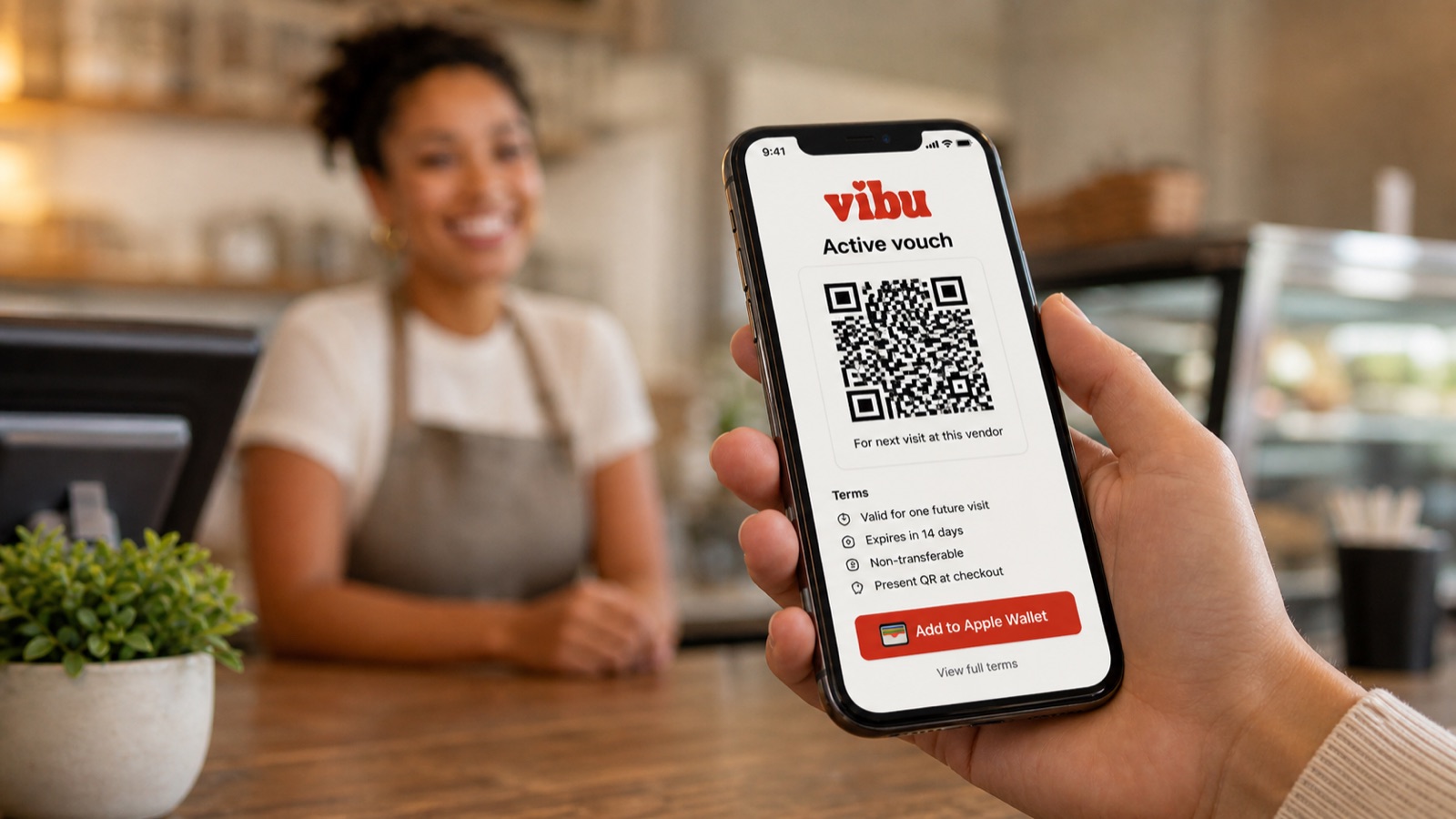 A customer's hand holds a phone showing the active Vibu voucher: a QR code, terms below, and Add to Apple Wallet and Google Wallet buttons. The vendor is in soft focus behind the phone.